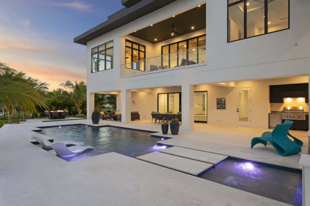 Modern rectangular pool with sun shelf and lounge chairs behind a two-story home at dusk, built by Curtis Pools