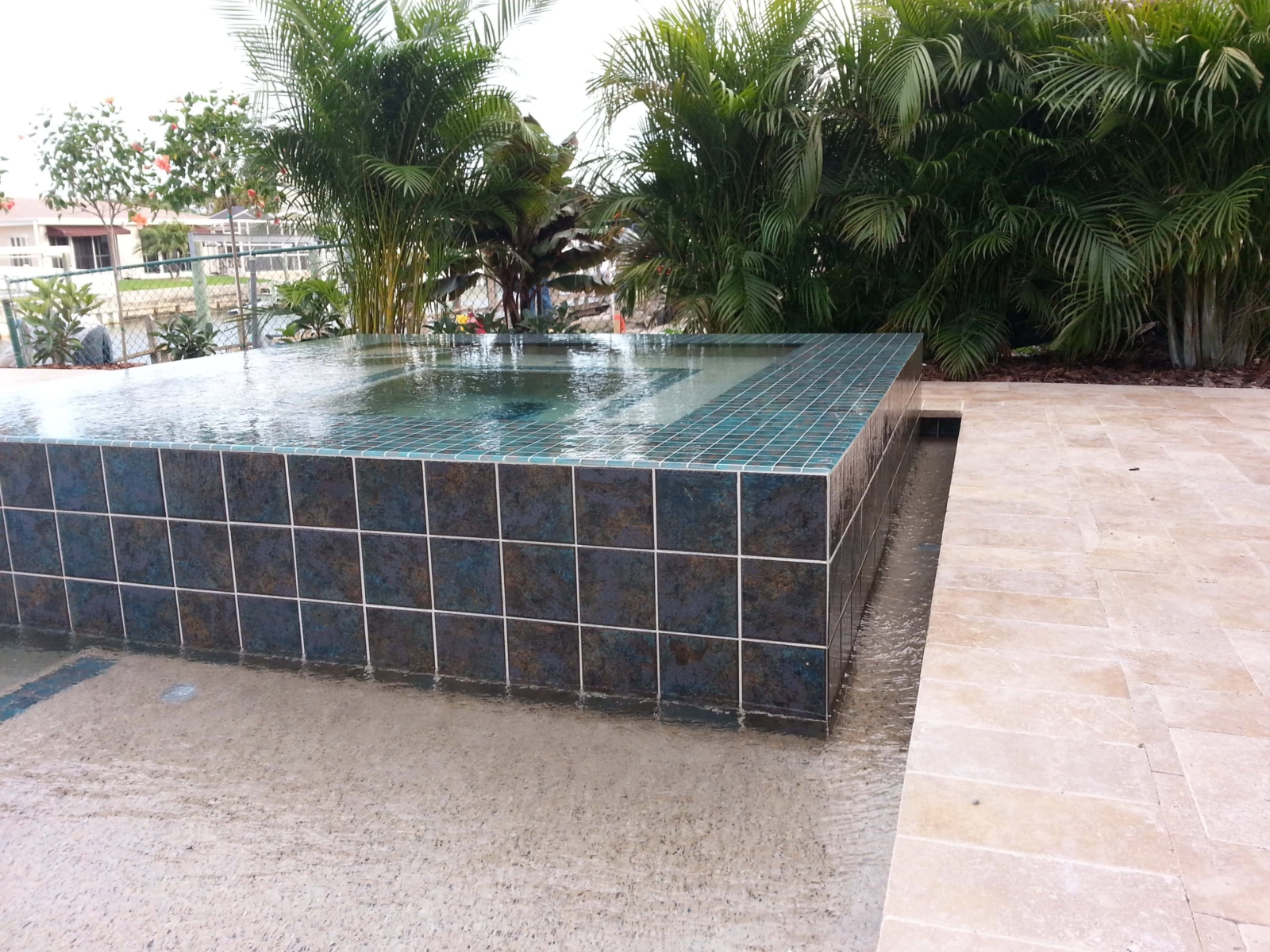 Raised square spa with blue tile overlooking a canal, palm trees and paver deck